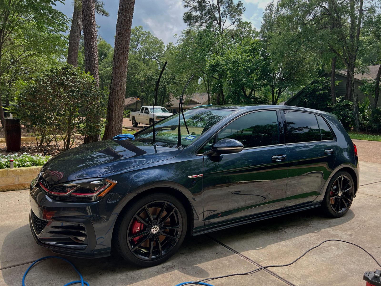 Dark gray Volkswagen hatchback with raised wipers in a tree-lined driveway